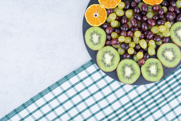 A dark plate full of grapes, kiwi and orange on tablecloth