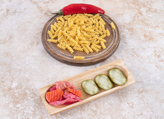 Assorted pickle tray next to a board with chili pepper and raw pasta pile on marble background