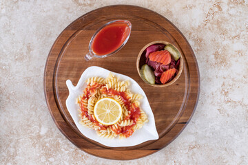 Wooden tray under a platter of cooked pasta, ketchup and a bowl mixed pickles on marble background