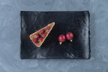 Cake slice decorated with raspberries on a black platter with two decorative balls on marble background