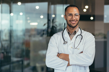 Portrait of young happy and smiling doctor, man in medical coat and stethoscope smiling and looking at camera, Arab doctor with crossed arms working inside modern clinic office. - Powered by Adobe
