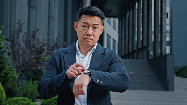 Angry worried 40s Asian businessman male frustrated boss manager CEO waiting meeting outdoors in city nervous annoyance frustrated pointing on wristwatch late time lateness deadline punctual wait
