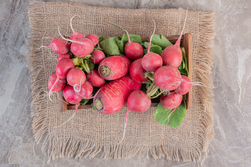 A wooden bowl full of turnips on turnip leaves sitting on a piece of fabric on marble background