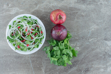 Fresh cilantro, onion and pomegranate salad next to a bundle of cilantro, red onion and a single pomegranate on marble background