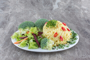 Portion of pilau garnished by chopped pepper, cabbage, greens, carrot and broccolie pieces on a platter on marble background