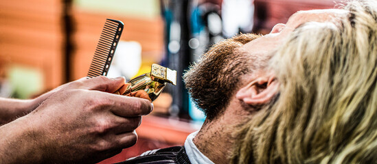 Bearded man in barbershop. Man visiting hairstylist in barbershop. Barber works with a beard clipper. Hipster client getting haircut. Hands of a hairdresser with a beard clipper, closeup - obrazy, fototapety, plakaty