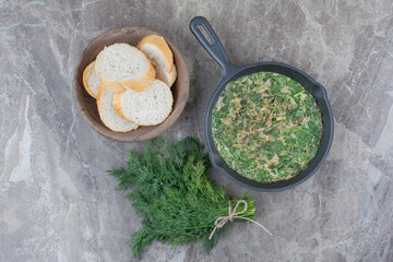 A dark pan of fried eggs with greens and white bread on marble background