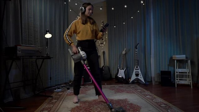 A beautiful girl in headphones cleaning the vacuum cleaner room and view at smartphone . Woman in a yellow sweater and hand hoover cleans the room