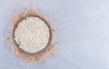 Wooden bowl placed on a pile of straw and filled with oat on marble background