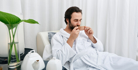 Portrait of handsome man in white bathrobe drinking healing tea while relaxing in wellness center. Men spa program