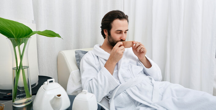 Portrait of handsome man in white bathrobe drinking healing tea while relaxing in wellness center. Men spa program