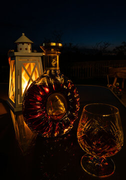 Glowing ring-shaped bottle of Remy Martin XO champagne cognac, and crystal brandy glass, on an outside table, in the dark, at night, backlit by candlelight from a lantern.
