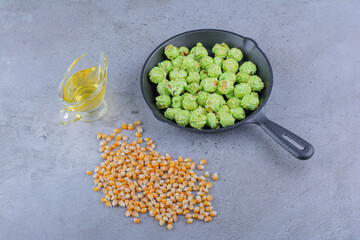 Small amount of corn kernel, glass of oil and a frying pan with candied popcorn on marble background