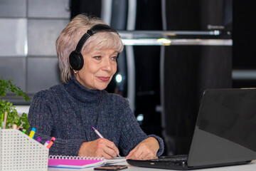 An elderly woman in her 60s and 65s is sitting in front of her laptop with headphones on. Concept:...