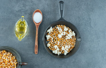 A jug and a pan full of corn grains, with a spoonful of salt and a glass of oil on marble background