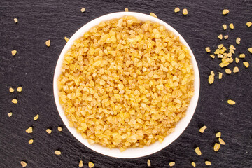 Raw bulgur grains in a white dish on slate stone, macro, top view.