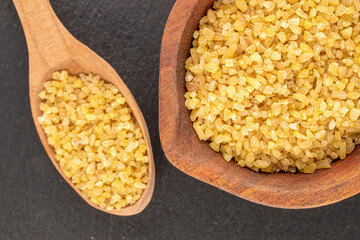 Raw bulgur grains in wooden cup with spoon on slate stone, macro, top view.