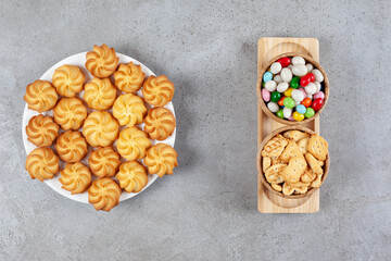 A full plate of homemade cookies with two bowls of crackers and candies on a tray on marble background