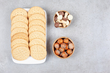 Two rows of biscuits on a platter next to small bowls of chocolate mushrooms and hazelnuts on marble background