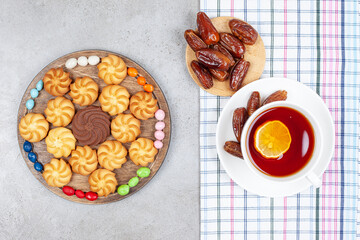 A cup of tea on a towel with dates and a wooden tray of arranged cookies and candy on marble background