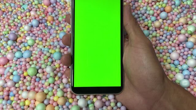 Chroma key green screen on smartphone vertical in male hand with colourful pastel rainbow foam ball beads in background
