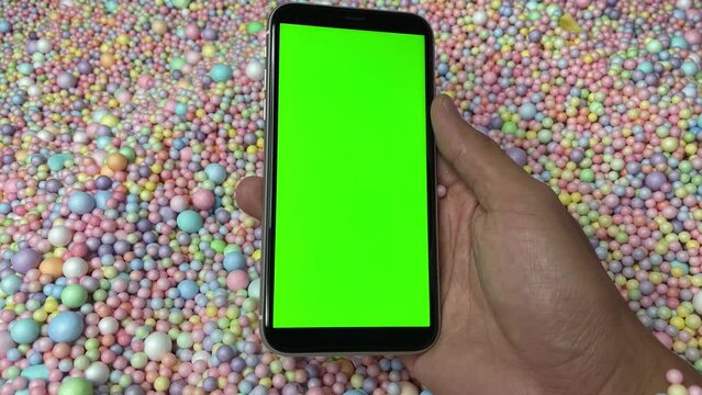Chroma key green screen on smartphone vertical in male hand with colourful pastel rainbow foam ball beads in background