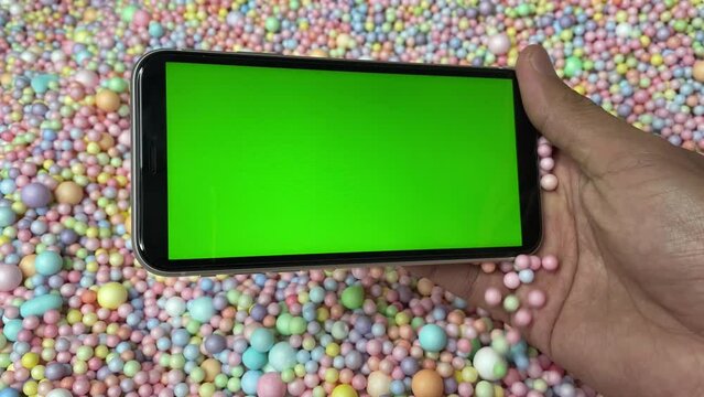Chroma key green screen on smartphone landscape in male hand with colourful pastel rainbow foam ball beads in background