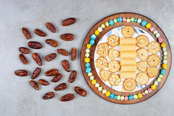 A plate of biscuits with a ring of candies on a wooden tray next to scattered dates on marble background