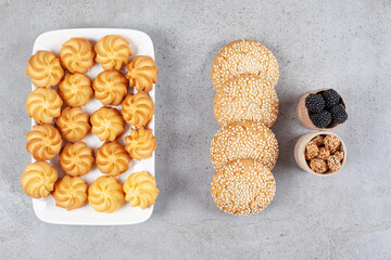 Small bowls of mullberries and glazed peanuts next to cookies on a plate and on marble background