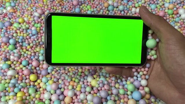 Chroma key green screen on smartphone landscape in male hand with colourful pastel rainbow foam ball beads in background