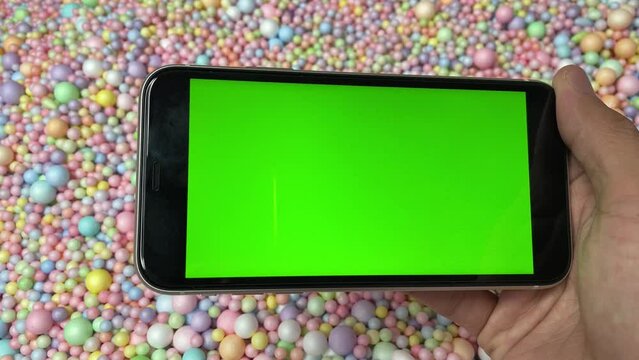 Chroma key green screen on smartphone landscape in male hand with colourful pastel rainbow foam ball beads in background