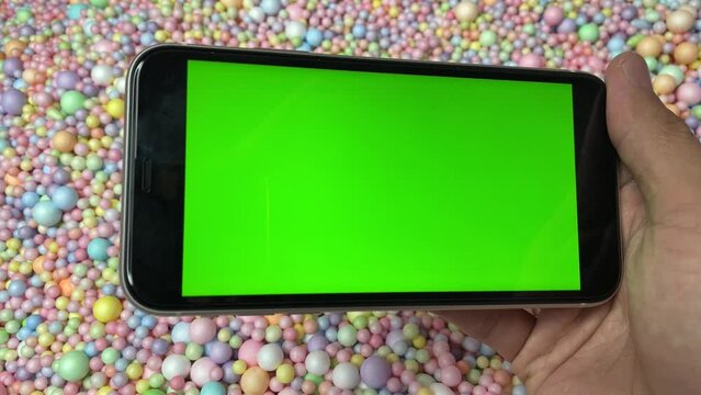 Chroma key green screen on smartphone landscape in male hand with colourful pastel rainbow foam ball beads in background