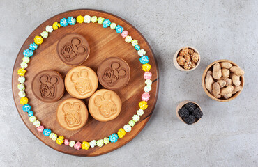 Cookies and a circle of candies on a wooden board next to bowls of peanuts and mullberries on marble background