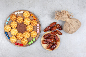 A bundle of dates and a sack with a wooden board of decoratively arranged candies and cookies on marble background