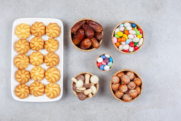Bowls of candies, hazelnuts, dates and chocolate mushrooms next to cookies on a plate on marble background
