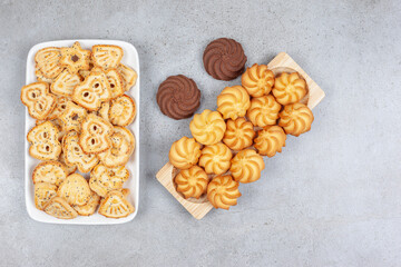 A wooden tray of cookies next to a plate of biscuit chips and two brown cookies in the middle on marble background