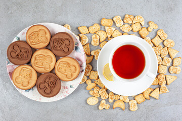A plate of tart cookies and a cup tea on saucer surrounded by biscuit chips on marble background