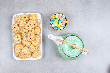 Plate of biscuits next to ornate teapot and bowl of candies on marble background