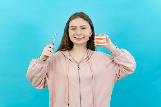 Front view half-body shot image of a young Caucasian teenage girl standing holding a dental instruments and a dental prosthesis teeth model while smiling at camera isolated on turquoise background.