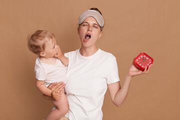 Indoor shot of young mother with bun hairstyle holding her toddler daughter and red alarm clock, being sleepy, yawning, standing with closed eyes and with blindfold, isolated over brown background.