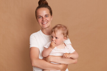 Image of smiling happy joyful woman with bun hairstyle posing with her toddler kid, looking at camera while holding her baby daughter isolated over brown background.
