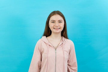 Front view half-body image of a happy young long-haired Caucasian teenage girl in a pink long sleeve hoodie, standing showing a beautiful smile with teeth to the camera on a turquoise background.