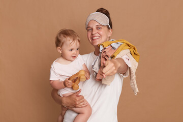 Portrait of happy positive woman wearing white t shirt and sleeping mask, mother with baby daughter holding laundry on shoulder, pointing finger to camera, isolated over brown background.