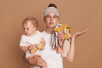 Portrait of sad upset stressed young mother with bun hairstyle holding her toddler daughter and clothing on her shoulder, spreading palm aside, posing isolated over brown background.