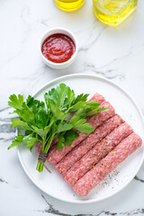 Raw serbian cevapi or cevapcici sausages with fresh parsley, high angle view on a white marble background, vertical shot