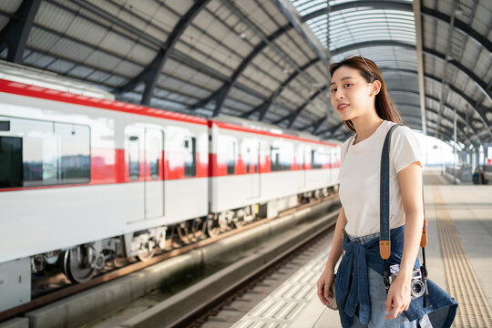 Selective focus of a happy Asian female traveler carrying a camera standing waiting for an arrival of skytrain on a platform with a blurred background of train in opposite side at a skytrain station.