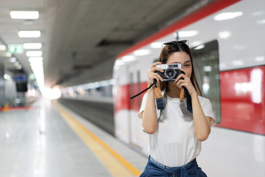 Selective focus, half-body image of a young beautiful Asian woman standing on a subway platform near a train, using a vintage film camera to take a picture in front of her at a blurred subway station.