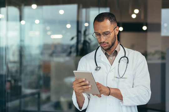 Serious and focused thinking african american doctor using tablet computer, man working inside modern clinic office