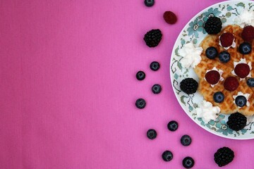 Waffle with cream and berries on a plate on a pink background.