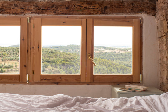 Habitación con vistas a la montaña, ventanas con vistas de naturaleza, habitación rústica con cama y nórdico blanco, en la mesita libros de bolsillo y movil
